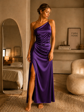 Woman in a purple one-shoulder dress standing in a room with a mirror and furniture.