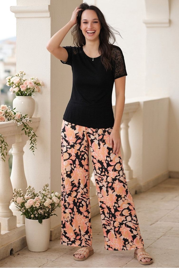 Woman wearing a black top and floral pants standing on a balcony with flowers and columns.
